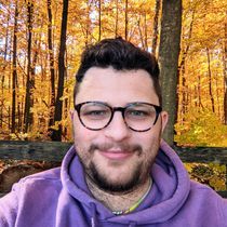 Simon is wearing a purple sweatshirt and glasses in a forest. 