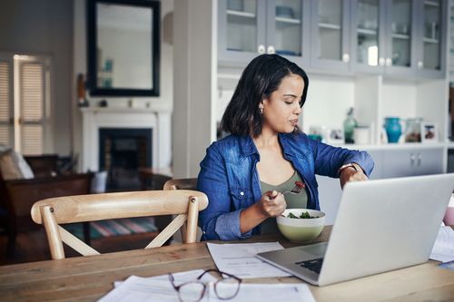 A woman sits at a computer and looks at her watch as she eats food