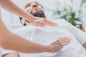 Man in a white t-shirt receiving a reiki healing session. He is lying down and the practitioner is moving her hands over his torso.