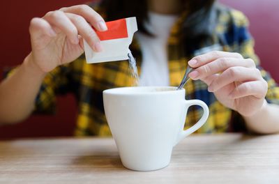 Woman wearing a yellow and black plaid shirt is pouring a sweetener packet into a white mug.