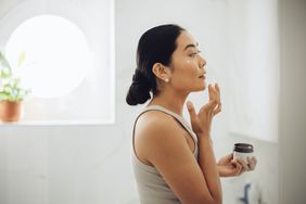 An Asian woman is wearing a tan tank top and has her hair pulled back. She is applying a cream to her face in the bathroom. 