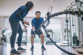 Asian Malay female personal trainer encouraging Asian Malay man practicing challenging battle rope in gym