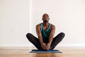 Black man sitting in butterfly pose on a yoga mat in a room with a white wall and hardwood floors