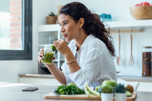 Women in her kitchen drinking a green juice with other greens in front of her