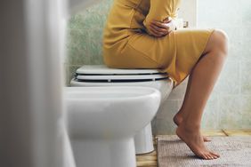 Woman in a yellow dress sitting on top of a toilet clutching her stomach