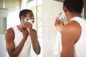 Young black man wearing white tank top is standing in front of bathroom mirror shaving his face.