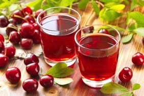 two glasses of cherry juices on a wood table with whole cherries around them
