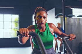 Black woman in green tank top with determined, concentrated face, training on stationary bike and sweating.. 