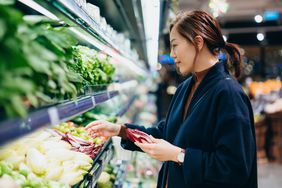 Asian woman with ponytail and navy jacket chooses from vegetables at a grocery store.