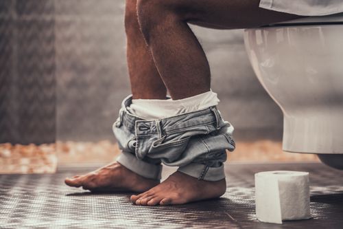 A person has their jeans around their ankles as they sit on the toilet with a roll of toilet paper next to them