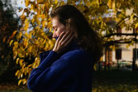 A women pulls her sweater over her mouth with her eyes closed outside during Fall.
