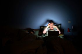 A man sits in bed with a laptop at night, next to a sleeping woman.