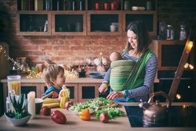 Happy young family, beautiful mother with two children, adorable preschool boy and baby in sling cooking together in a sunny kitchen